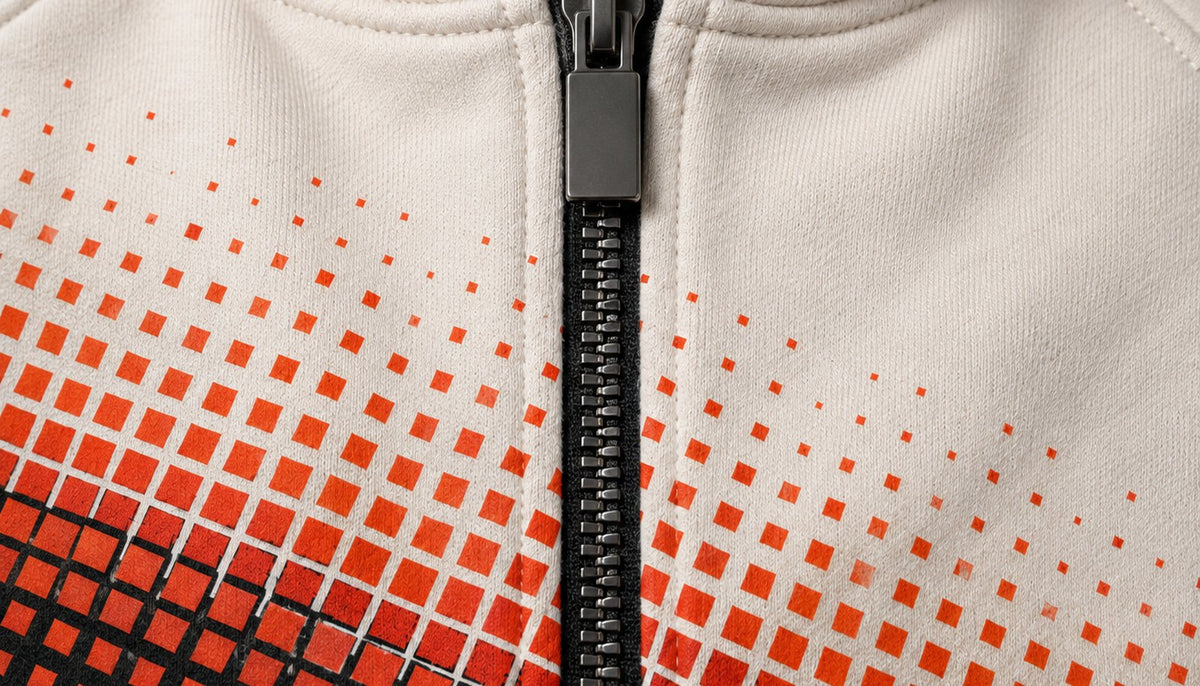 Gros plan sur le zip central : curseur et tirette métalliques sombres, ruban noir, et le dégradé de carrés orange qui se coupe sur la ligne du zip.